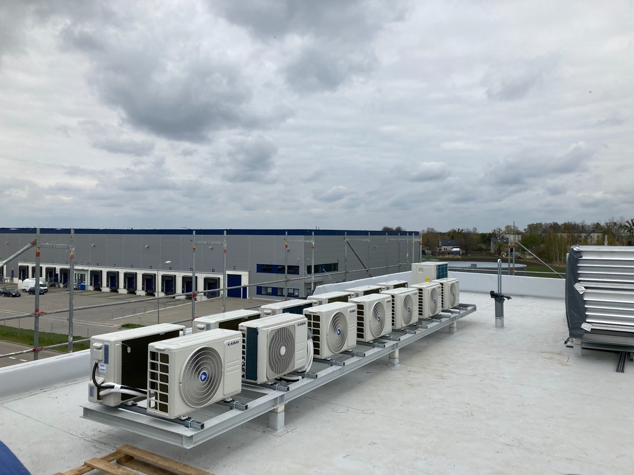 Rząd białych jednostek zewnętrznych klimatyzacji marki Kaisai, zamontowanych na metalowej konstrukcji na dachu budynku komercyjnego z rampami załadunkowymi w tle, pochmurne niebo.