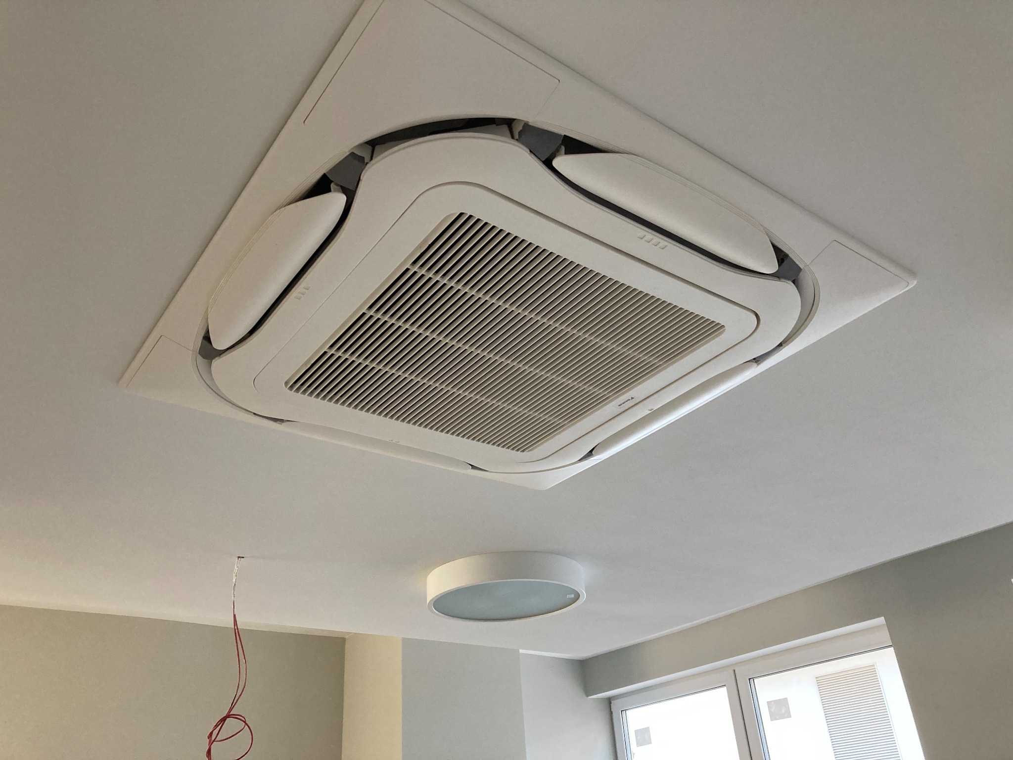 Kasetonowa jednostka klimatyzacji sufitowej w jasnym pomieszczeniu, widoczne wystające przewody elektryczne, okrągła lampa sufitowa w tle.