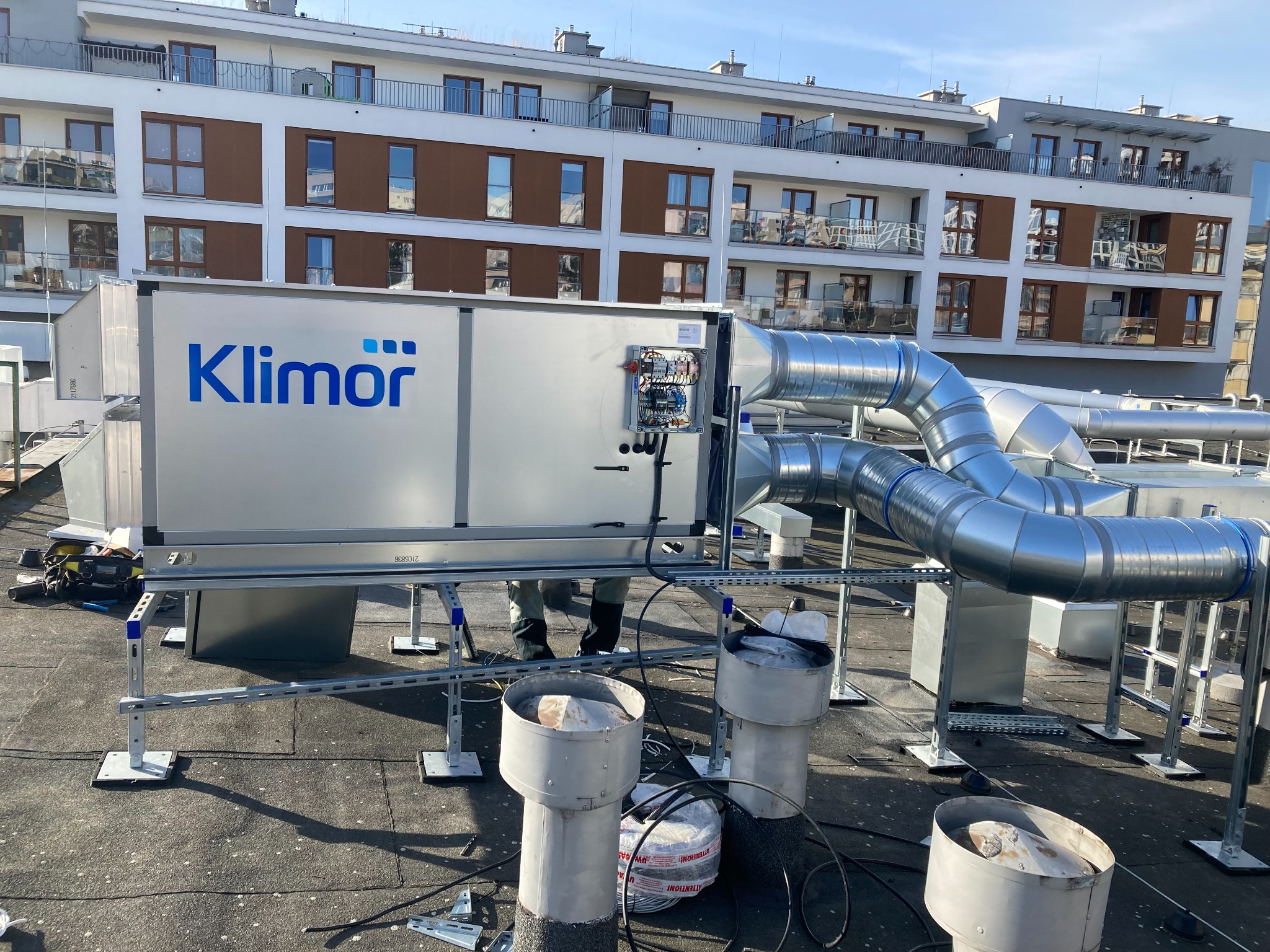 Instalacja centrali wentylacyjnej Klimor na dachu budynku wielorodzinnego, widoczne przewody wentylacyjne, elementy montażowe i częściowo sylwetka instalatora.