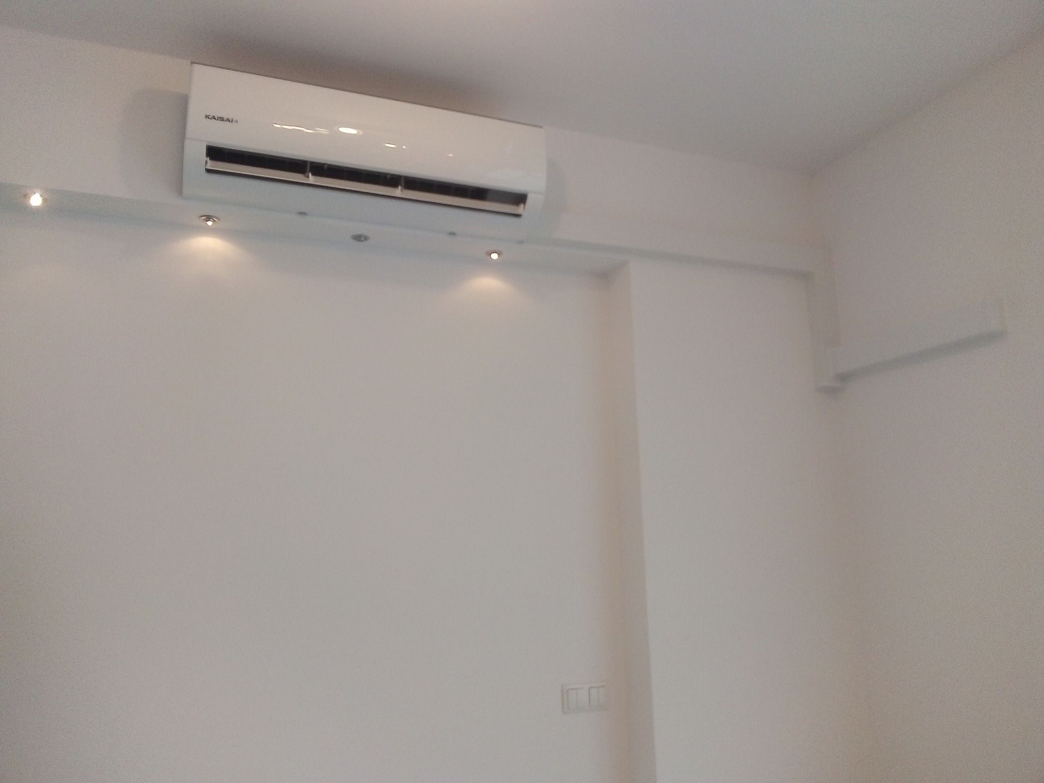 Biały klimatyzator marki Kaisai zamontowany na ścianie w pomieszczeniu z oświetleniem punktowym.