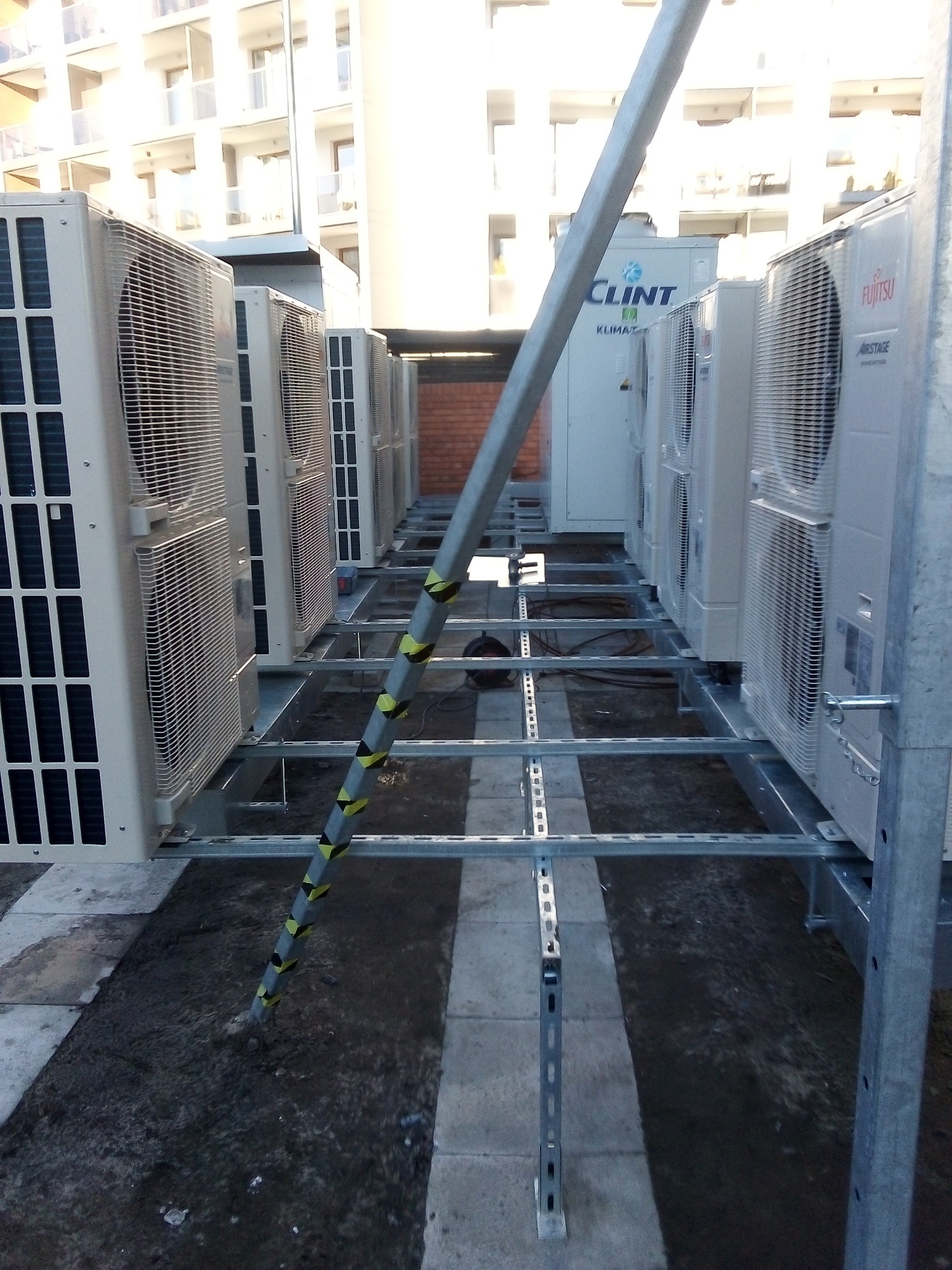 Rząd zewnętrznych jednostek klimatyzacyjnych Fujitsu i Clint na metalowej konstrukcji wsporczej, widok z dołu, budynek w tle.