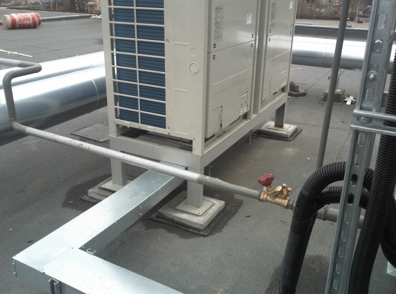 Dwa moduły klimatyzacji przemysłowej na dachu budynku, połączone rurami i zaworami, z widocznymi elementami instalacji wentylacyjnej i kanalizacyjnej.