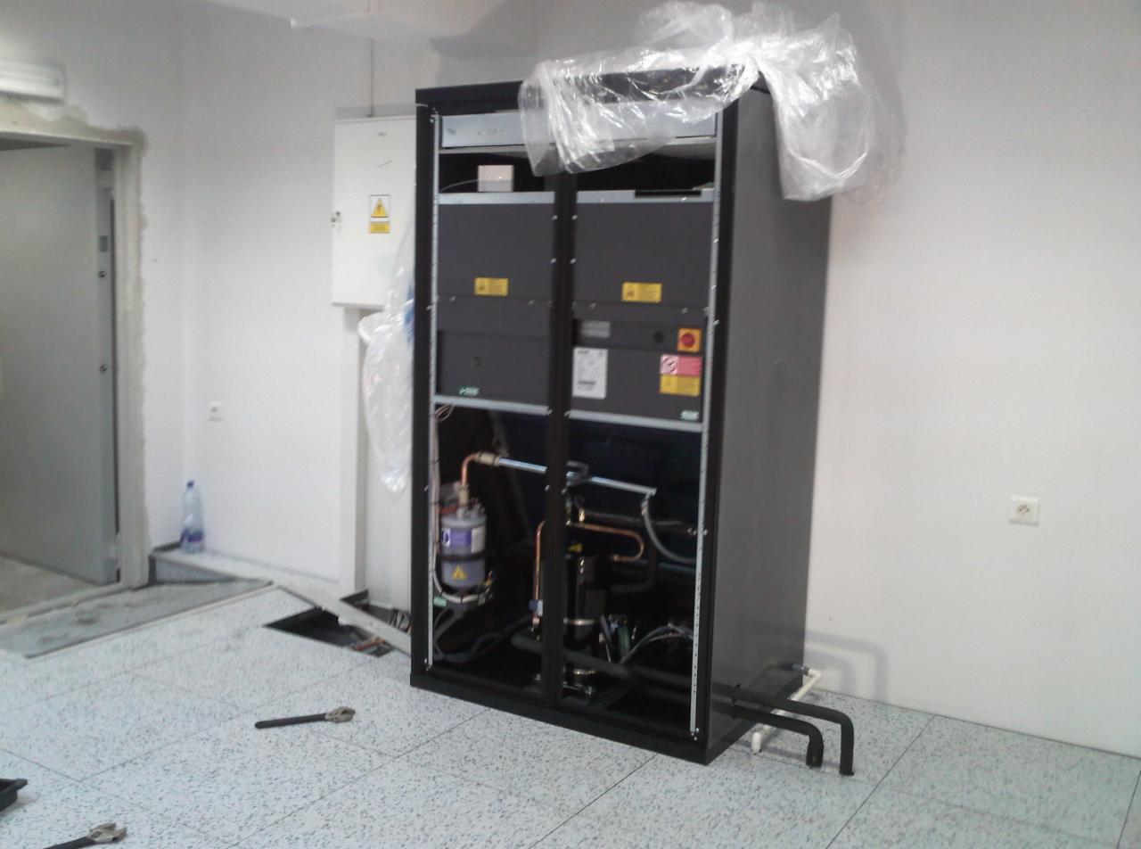 Duża, czarna centrala wentylacyjna w trakcie montażu, z widocznymi elementami instalacji, rurami miedzianymi i narzędziami na podłodze, zabezpieczona folią ochronną, w pomieszczeniu z białą ścianą...