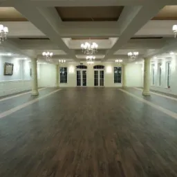 Duża sala z panelami podłogowymi w odcieniach brązu, ozdobnymi kolumnami i żyrandolami. Widok na przestronne wnętrze z symetrycznym układem okien i drzwi.