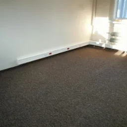 Ciemna wykładzina dywanowa w jasnym pomieszczeniu biurowym z białymi listwami przypodłogowymi i grzejnikami. W tle błękitny kask na parapecie okna.