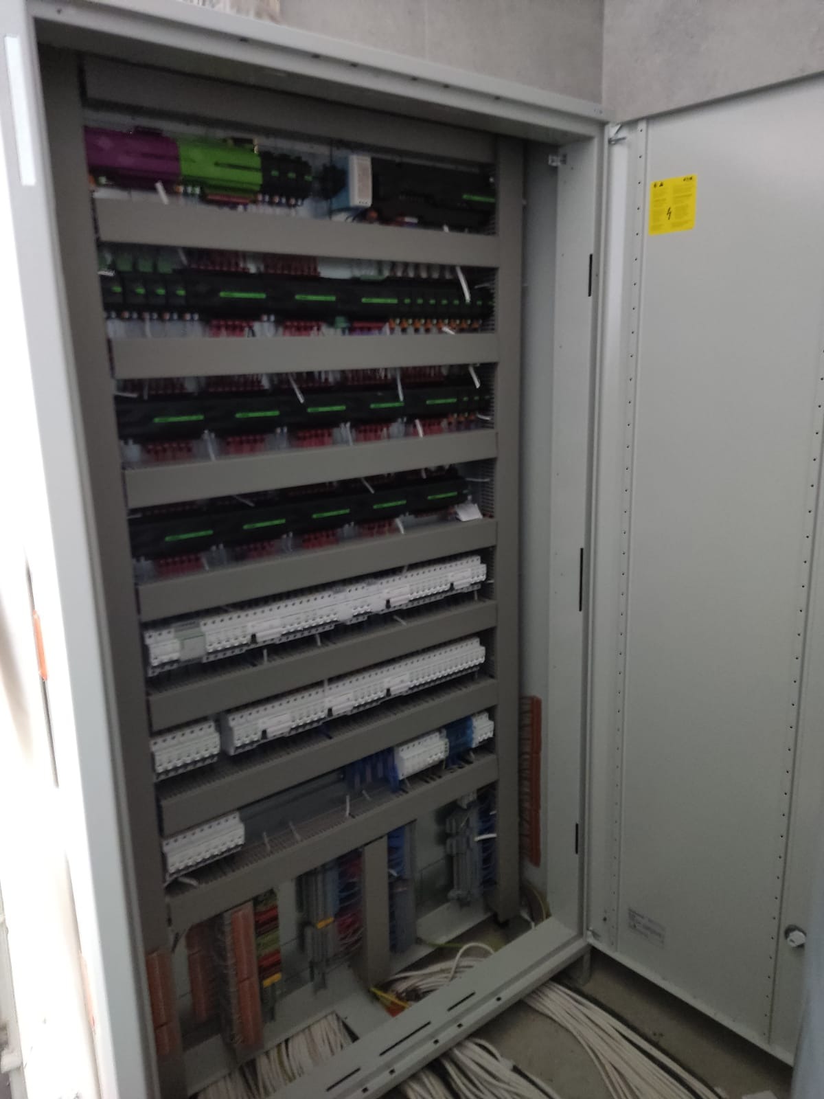 Otwarta szafa elektryczna z licznymi rzędami bezpieczników, przewodów i złączek, widoczne kable na podłodze.