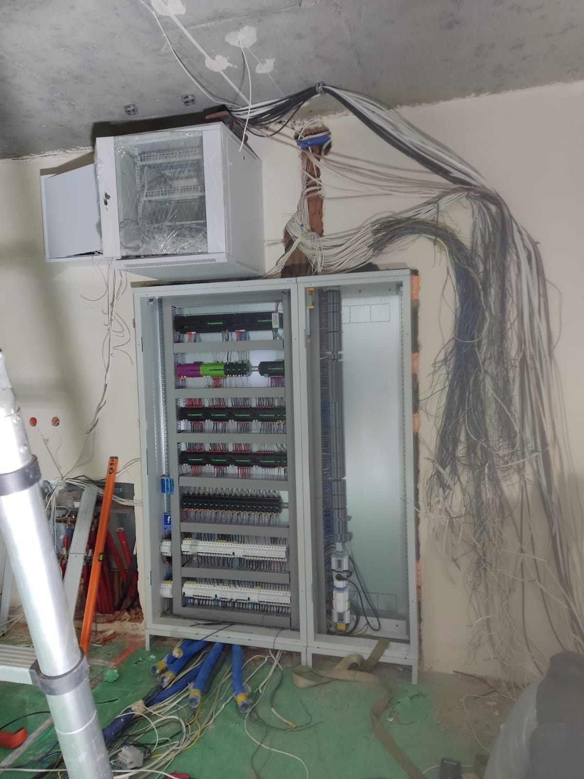 Rozdzielnia elektryczna w trakcie montażu, widoczne liczne przewody wychodzące ze ściany i sufitu, oraz szafka RACK z folią ochronną, częściowo otwarte drzwi rozdzielnicy, elementy instalacji...