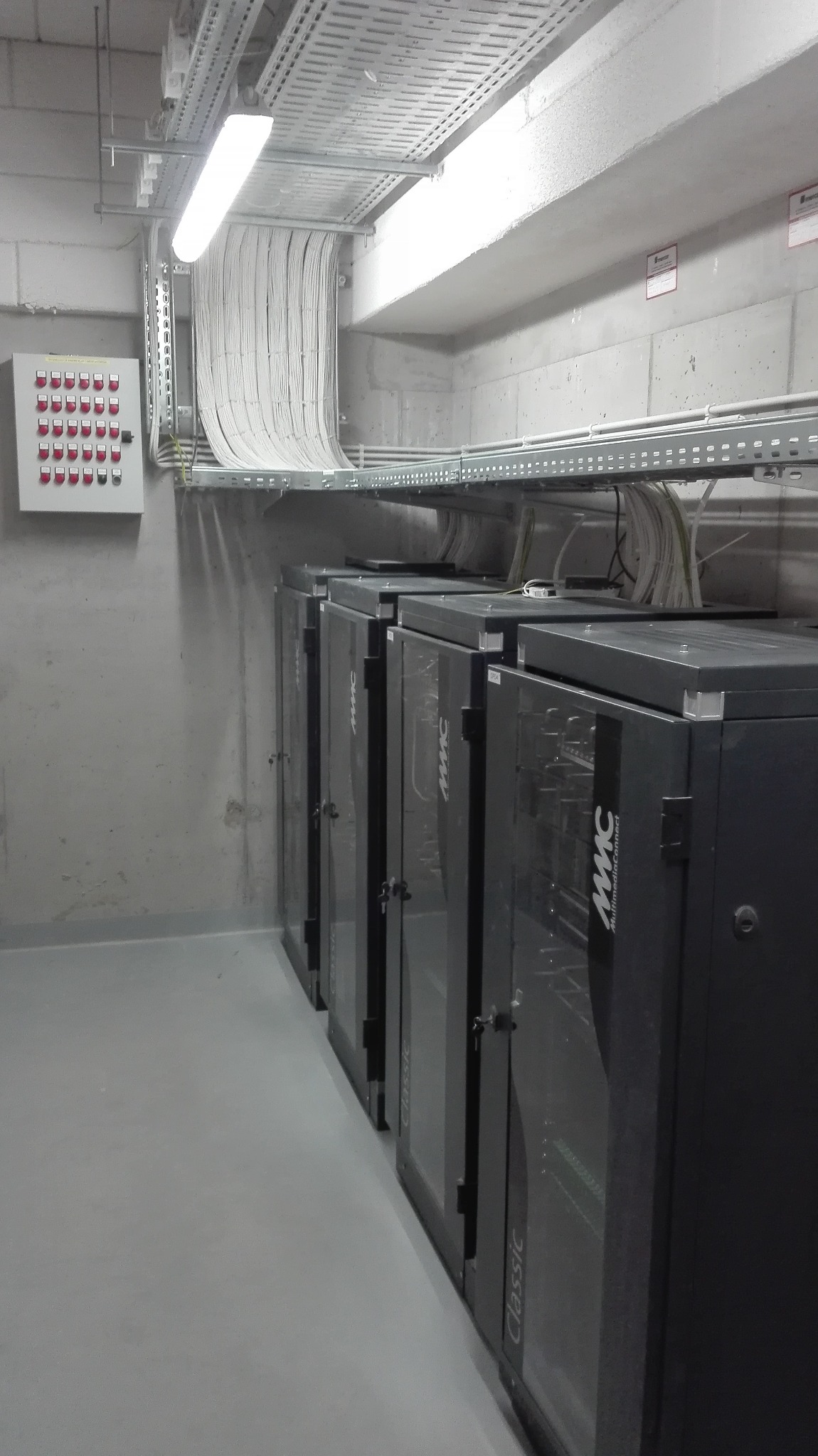 Rząd szaf serwerowych w szarym pomieszczeniu z betonowymi ścianami, z widocznymi kablami prowadzonymi pod sufitem na metalowych korytkach. Widoczna skrzynka z przyciskami na ścianie.