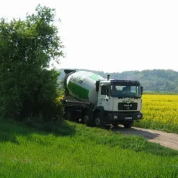 Ciężarówka betonomieszarki MAN na polnej drodze, z zielonymi akcentami na bębnie, na tle żółtego pola i drzew. Widok z bliska.