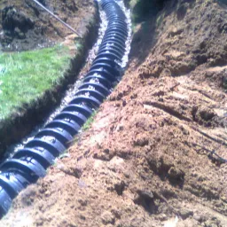 Instalacja drenażowa z czarnych rur karbowanych w wykopie, z kopcami ziemi po bokach. W tle widoczny zielony zbiornik i trawa.