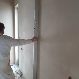 Pracownik w ubraniu roboczym sprawdza pion pionową poziomicą na świeżo nałożonym tynku na ścianie wewnątrz budynku, widoczne puszki elektryczne i przewody.