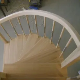 Drewniane schody kręcone z jasnego drewna z balustradą z okrągłych szczebelków i łukowatą poręczą, widok z góry.