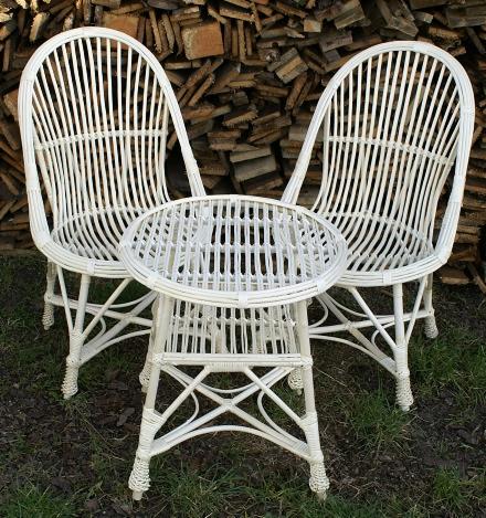 Biały, wiklinowy zestaw mebli ogrodowych: dwa krzesła i okrągły stolik, na tle stosu drewna i trawy. Klasyczny styl, naturalne materiały.