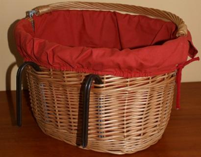 Wiklinowy kosz rowerowy z czerwoną wyściółką i metalowymi uchwytami, umieszczony na drewnianym blacie. Kosz pleciony z jasnego wikliny, widoczne detale splotu.
