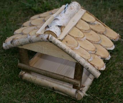 Drewniany karmnik dla ptaków z kory brzozowej, ręcznie robiony, na trawie. Dach pokryty plasterkami drewna, naturalne materiały, rustykalny styl.