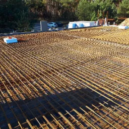 Siatka zbrojeniowa ułożona na płaskiej powierzchni, przygotowana do zalania betonem, widok z góry na tle lasu i częściowo ukończonej konstrukcji budynku.