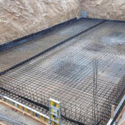 DOM-BET-BUD Wojciech Cichulski - Zbrojenie płyty fundamentowej w wykopie, widoczne szalunki i poziomica, częściowo zalane wodą.