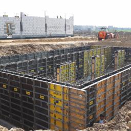 DOM-BET-BUD Wojciech Cichulski - Szalunek fundamentowy z widocznym zbrojeniem, przygotowany do zalania betonem, na placu budowy z budynkiem w stanie surowym otwartym w tle.