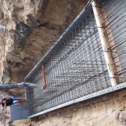 Wykop z betonową ścianą zbrojoną stalą, w trakcie wylewania betonu z pompy betonomieszarki, widoczny pracownik w niebieskich spodenkach.