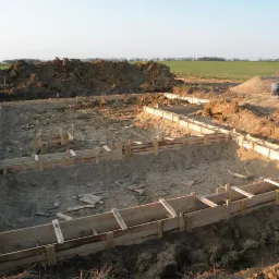 Szeroki kadr z ziemi ukazuje drewniane szalunki fundamentów pod budynek na tle pola i wzgórz. Widoczne są ślady opon i wykopana ziemia.