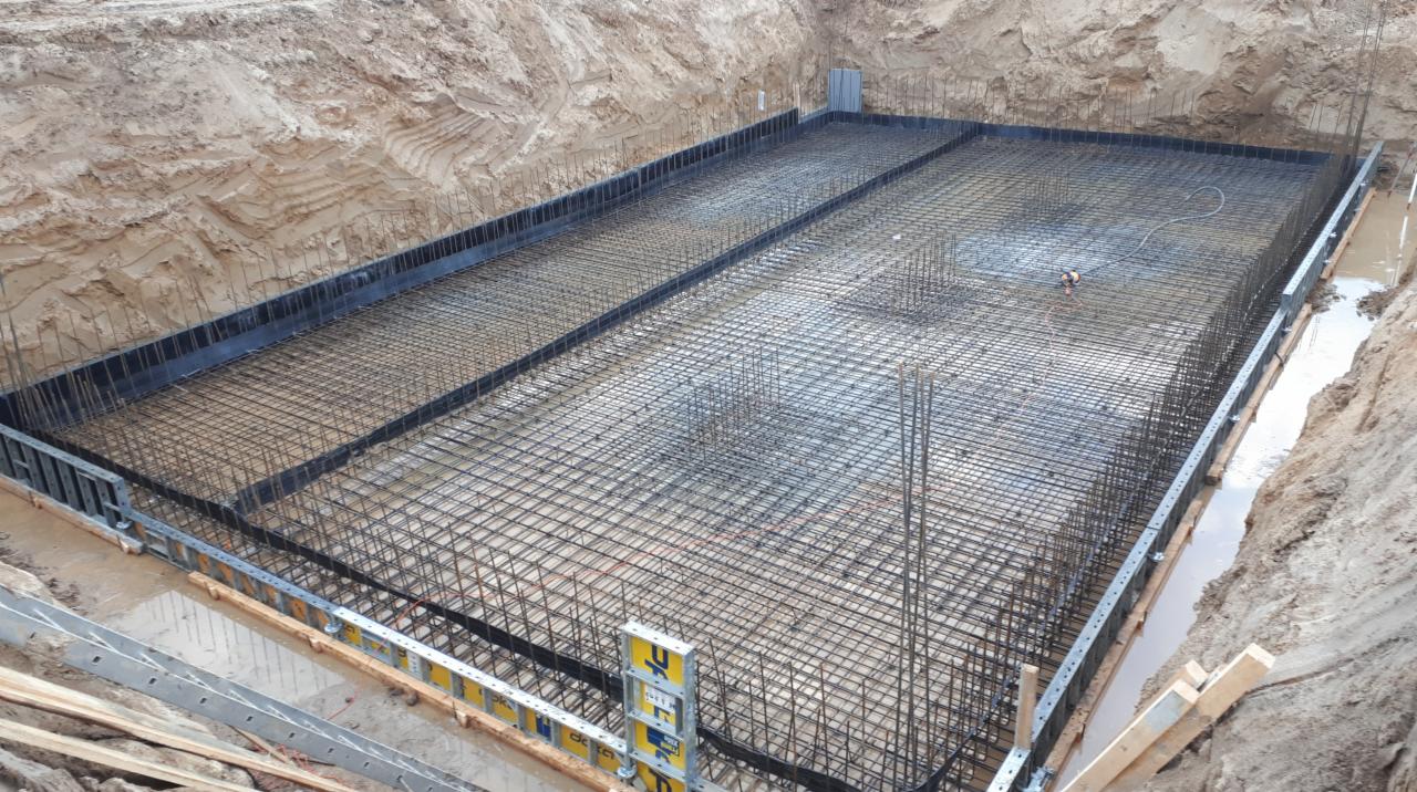 Zbrojenie płyty fundamentowej w wykopie, widoczne szalunki i poziomica, częściowo zalane wodą.