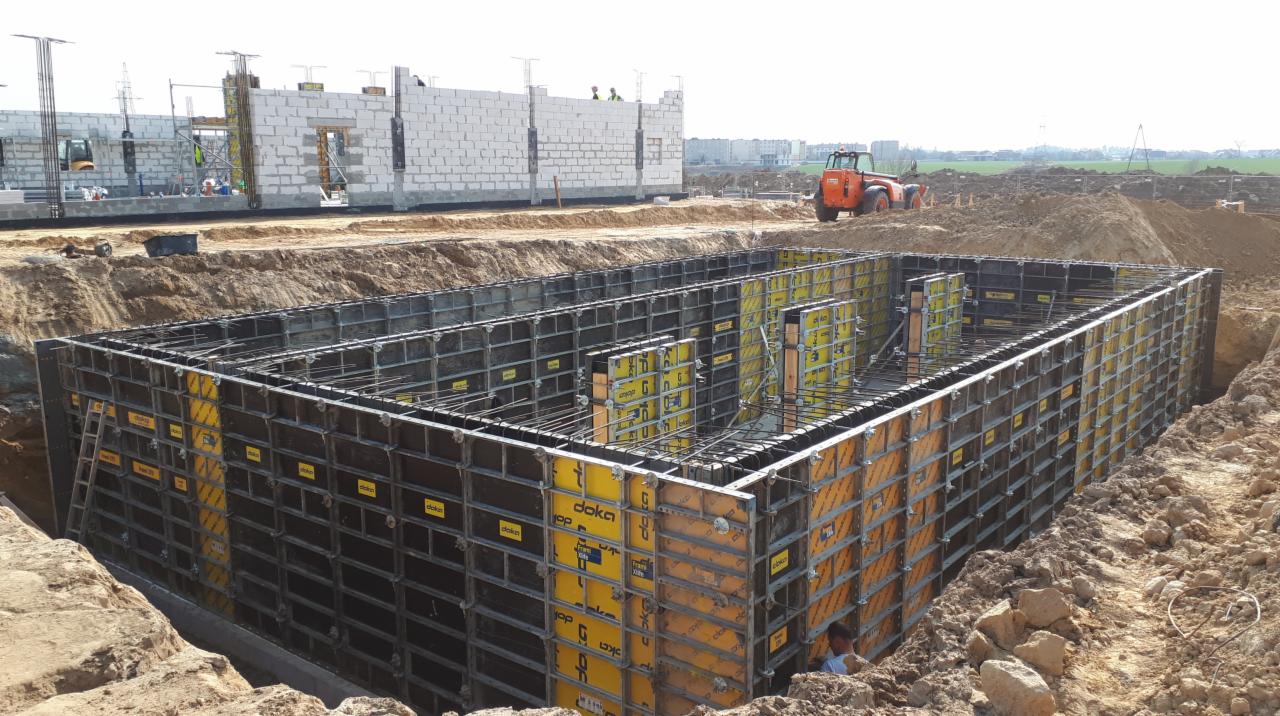 Szalunek fundamentowy z widocznym zbrojeniem, przygotowany do zalania betonem, na placu budowy z budynkiem w stanie surowym otwartym w tle.