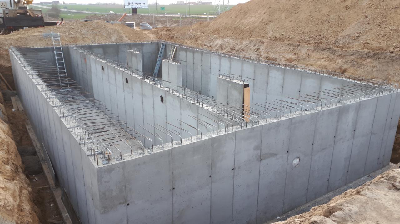 Żelbetowa konstrukcja w wykopie, widoczne wystające pręty zbrojeniowe, drabina oparta o ścianę, w tle billboard reklamowy.