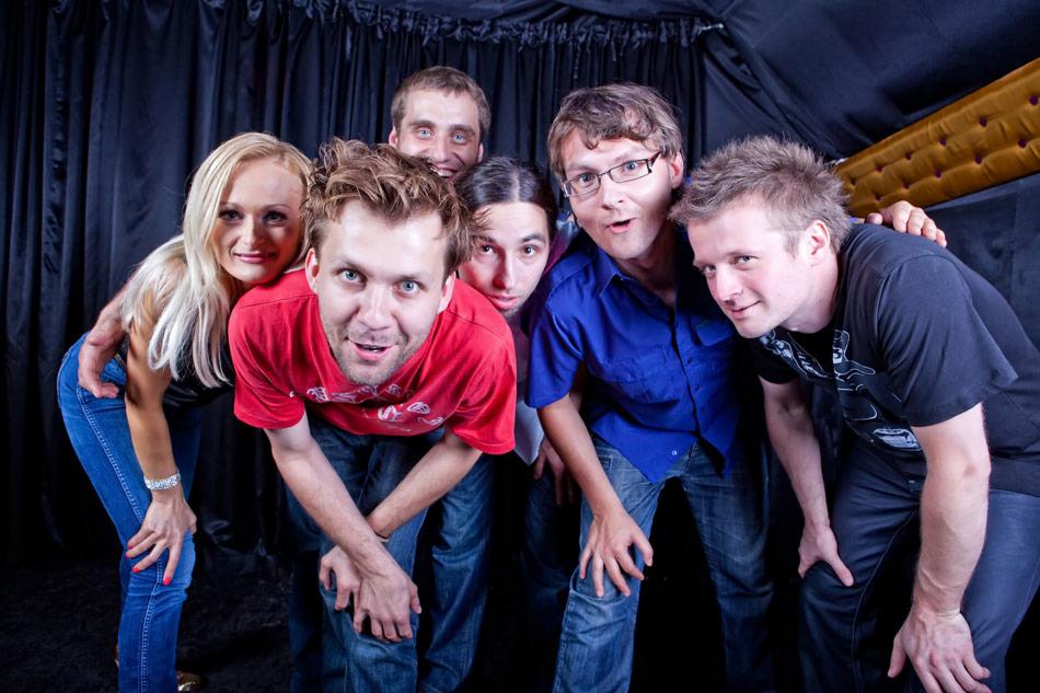 Sześcioosobowa grupa w dżinsach i koszulkach, pozująca z bliska na tle czarnej, drapowanej zasłony.