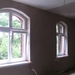 Wnętrze pokoju z dwoma oknami łukowymi w trakcie tynkowania ścian. Widoczne surowe tynki i wystające przewody. Okna białe, otwarte, za nimi zieleń.