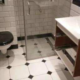 Wykończona łazienka z biało-czarną mozaiką na podłodze i ścianach, kabiną prysznicową ze szkła i umywalką z szafką w odcieniach brązu. Widok na toaletę z czarną deską.