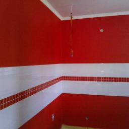 JAROSZY - Intensywnie czerwone ściany w pomieszczeniu z białymi płytkami i bordową mozaiką, widoczne przewody elektryczne na suficie.