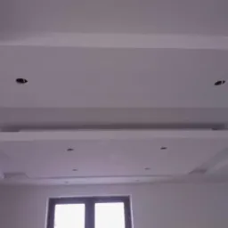 Biały, wielopoziomowy sufit podwieszany z wnękami na oświetlenie punktowe, widok na okno w dolnej części kadru.