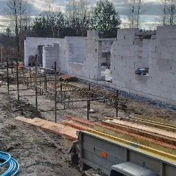 Wznoszenie ścian parterowego domu z białych bloczków, widoczne zbrojenie fundamentów i przyczepka z materiałami budowlanymi na placu budowy.