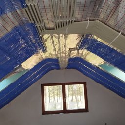 JAROSZY - Poddasze z oknem dachowym w trakcie prac wykończeniowych: widoczne rury instalacji grzewczej pod stropem, izolacja folią aluminiową i niebieską wełną mineralną na skosach, oraz surowa podłoga...