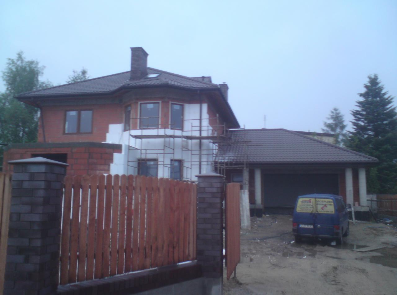 Dom w trakcie ocieplania, widoczne rusztowania i styropian. Częściowo wykończone ogrodzenie z ceglanymi słupkami. Na podjeździe zaparkowany niebieski bus.