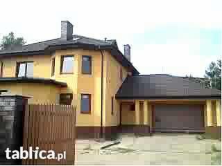 Żółty dom jednorodzinny z ciemną dachówką i garażem wbudowanym, widoczny fragment ogrodzenia i komin.