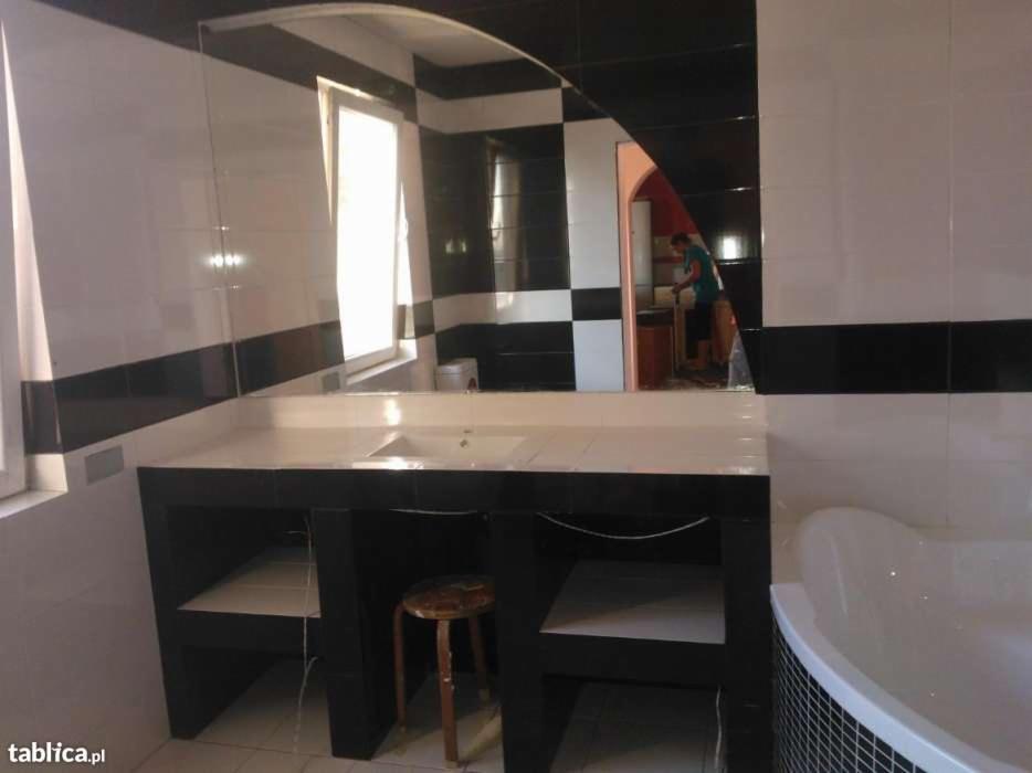 Wykończona łazienka z biało-czarnymi płytkami, dużym lustrem i zabudowaną umywalką z półkami, widoczna osoba w tle.