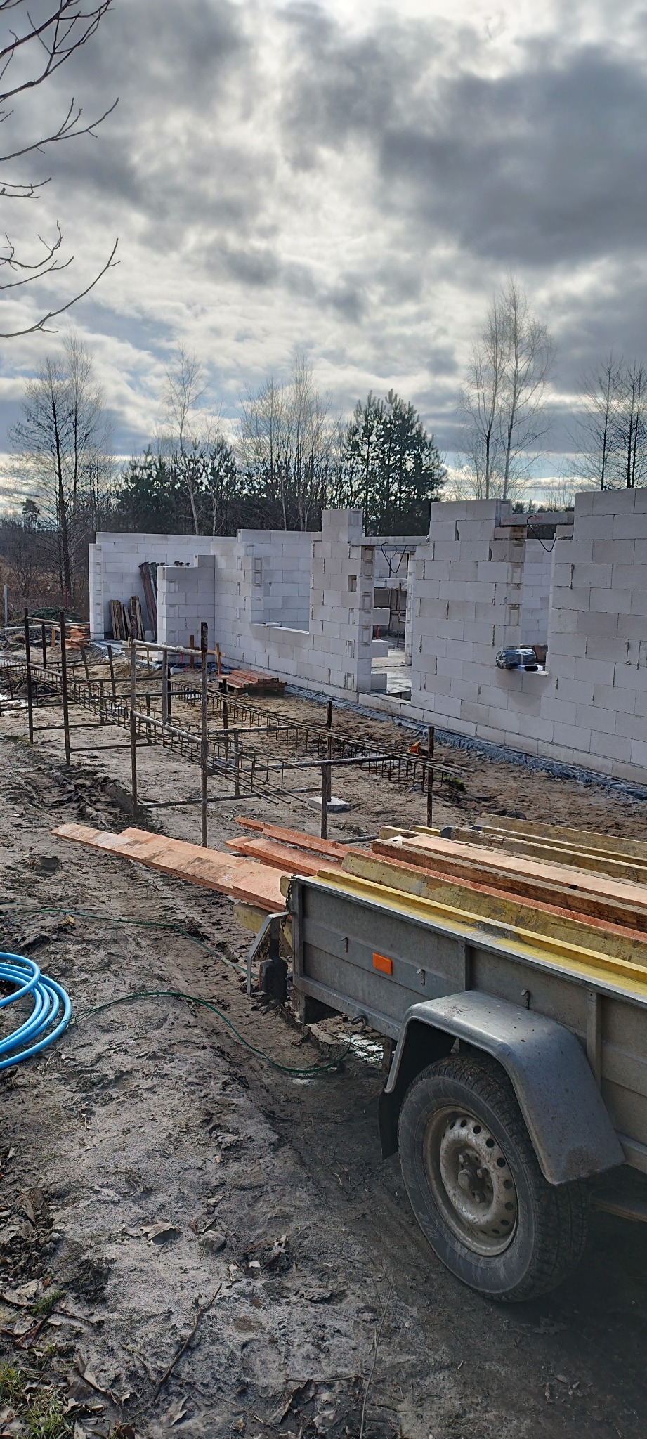 Wznoszenie ścian parterowego domu z białych bloczków, widoczne zbrojenie fundamentów i przyczepka z materiałami budowlanymi na placu budowy.