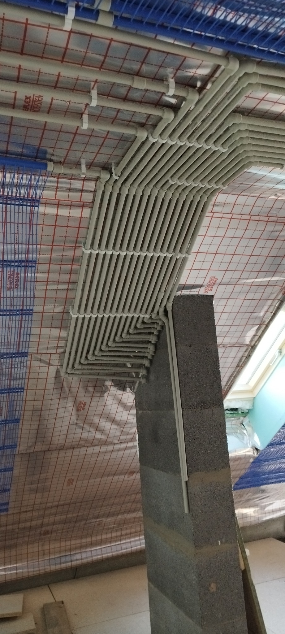 Równoległe rury instalacji pod sufitem przymocowane do ściany z bloczków betonowych, widoczne izolacja termiczna i fragment okna dachowego.