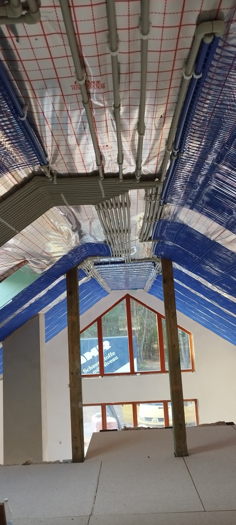 Instalacja ogrzewania podłogowego w poddaszu, widoczne rury grzewcze przymocowane do folii izolacyjnej z czerwonym nadrukiem, drewniane słupy konstrukcyjne oraz okno z brązową ramą.