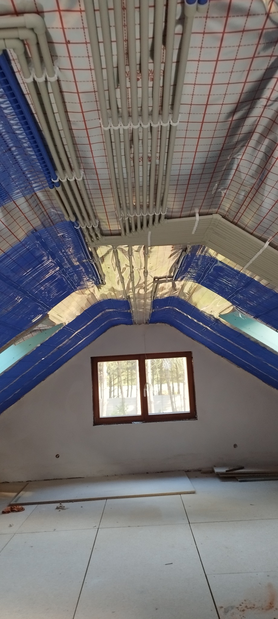 Poddasze z oknem dachowym w trakcie prac wykończeniowych: widoczne rury instalacji grzewczej pod stropem, izolacja folią aluminiową i niebieską wełną mineralną na skosach, oraz surowa podłoga...