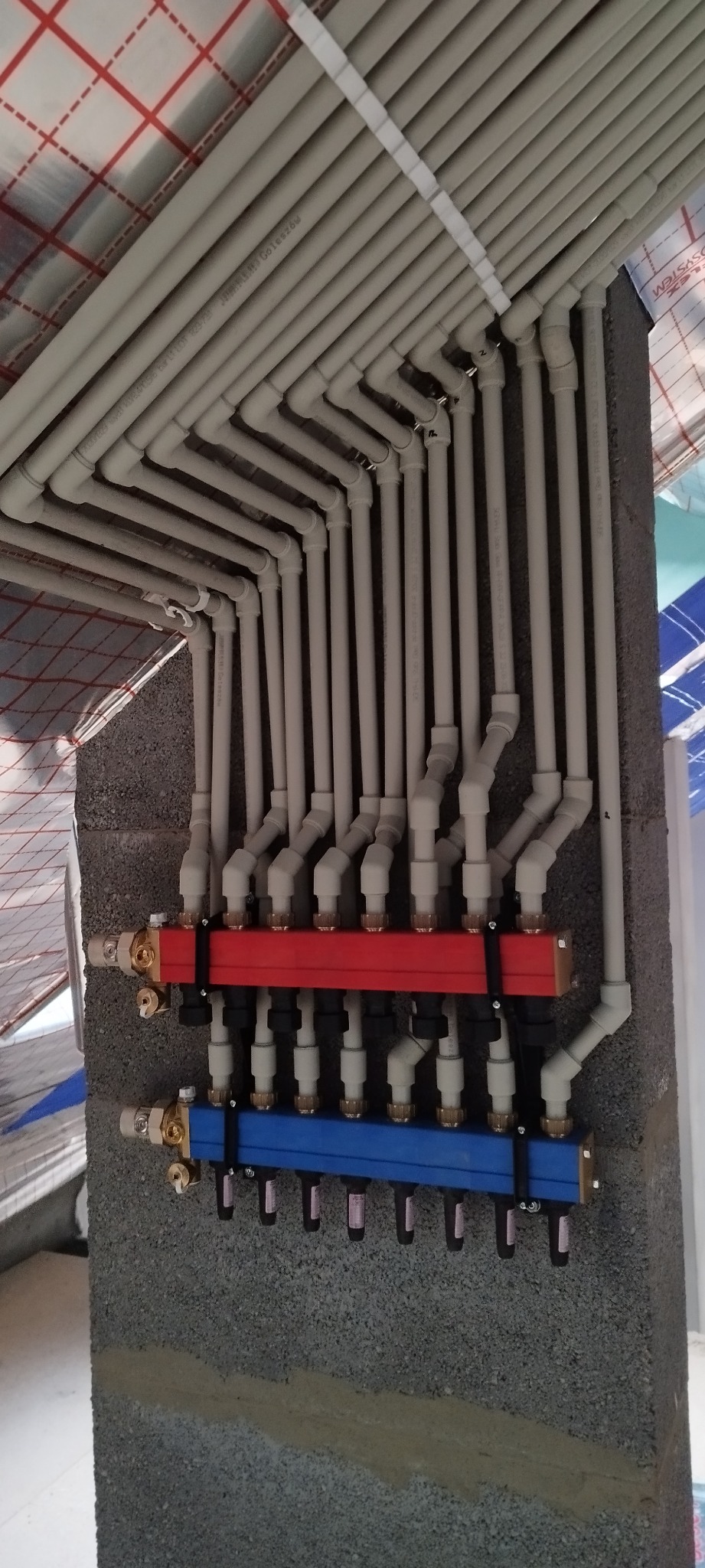 Rozdzielacz ogrzewania podłogowego z rurami PEX zamontowany na ścianie z betonu komórkowego, widoczne czerwone i niebieskie belki rozdzielcze.