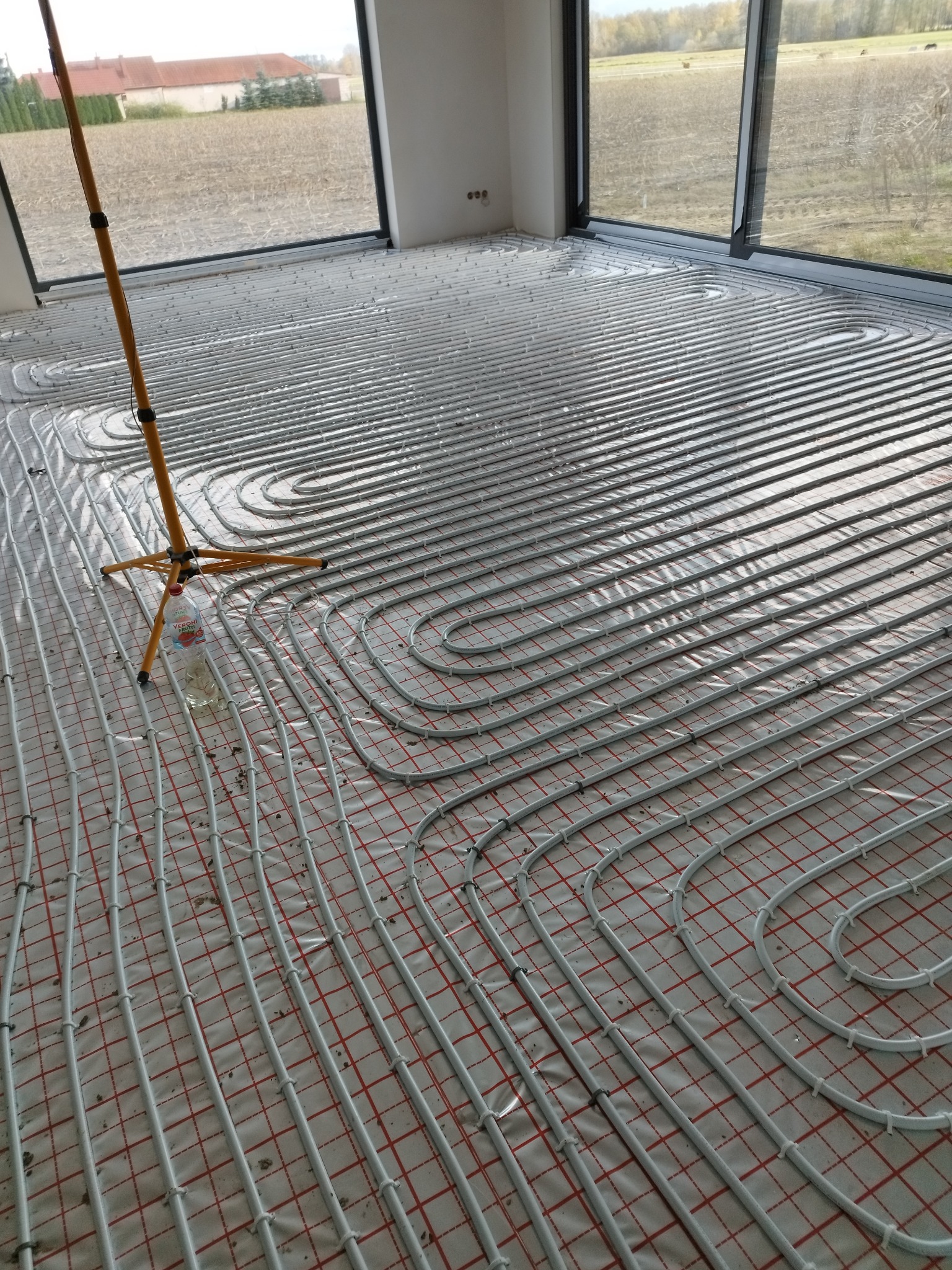 Rozłożone rury ogrzewania podłogowego na folii z czerwoną siatką, widoczna lampa budowlana i butelka wody, duże okna z widokiem na pole.