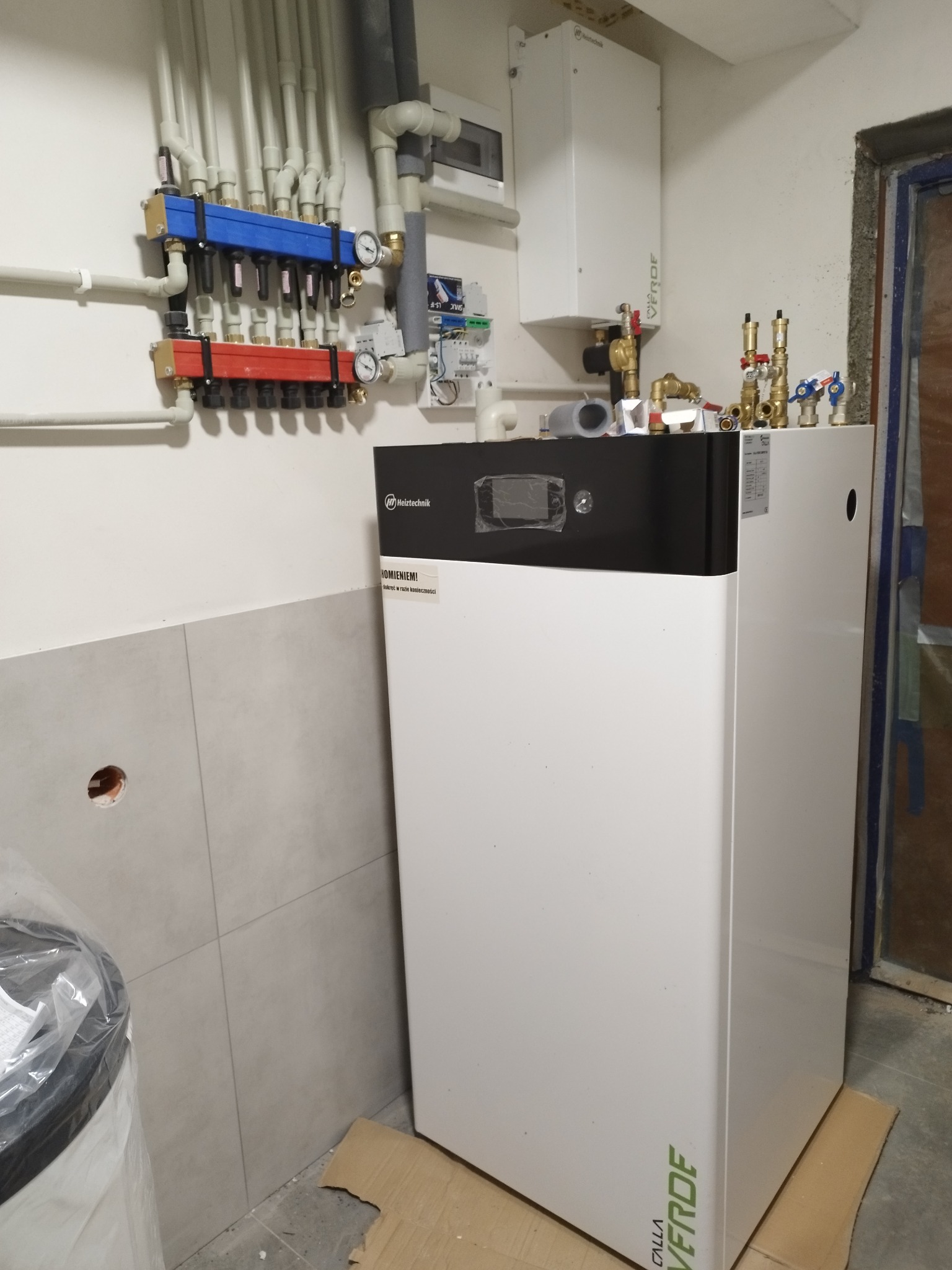 Nowoczesny piec grzewczy Heitztechnik z rozdzielaczem CO/CWU na ścianie, widoczne rury i armatura, szare płytki na ścianie w tle.