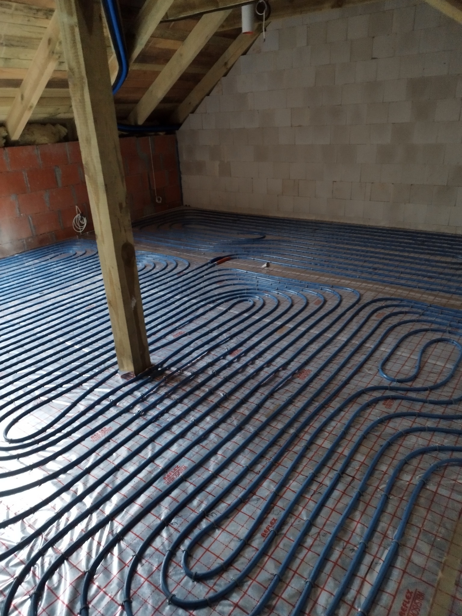 Niebieskie rury ogrzewania podłogowego ułożone na srebrnej folii refleksyjnej, widoczne elementy konstrukcji dachu i ściany z białego bloczka betonu komórkowego.