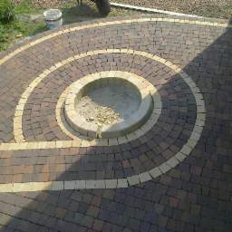 Okrągła kompozycja z kostki brukowej w odcieniach brązu i beżu, z centralnym okręgiem wykończonym kostką. Widok z góry, cień na kostce przy budynku.