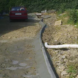 Formowanie krawężnika z betonu wzdłuż podjazdu, widoczny samochód osobowy i przygotowane podłoże. Prace brukarskie w toku, widok z perspektywy.