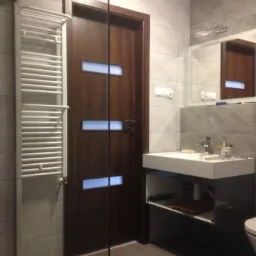 Wykończona łazienka z szarymi płytkami, brązowymi drzwiami z przeszkleniami i grzejnikiem drabinkowym. Nowoczesna umywalka z szafką i lustro.