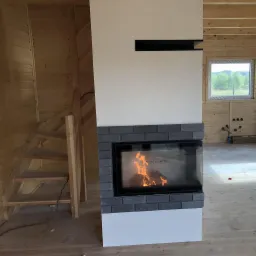 Wnętrze domu w trakcie budowy: kominek z szarej cegły z palącym się drewnem, drewniane ściany i schody, widok na zewnątrz przez okno.