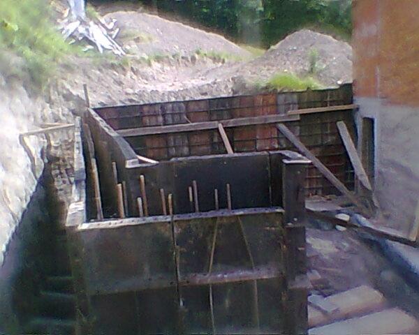 Szczegółowy widok na deskowanie fundamentów z wystającymi prętami zbrojeniowymi, przygotowane do zalewania betonem, otoczone ziemią i skarpą.